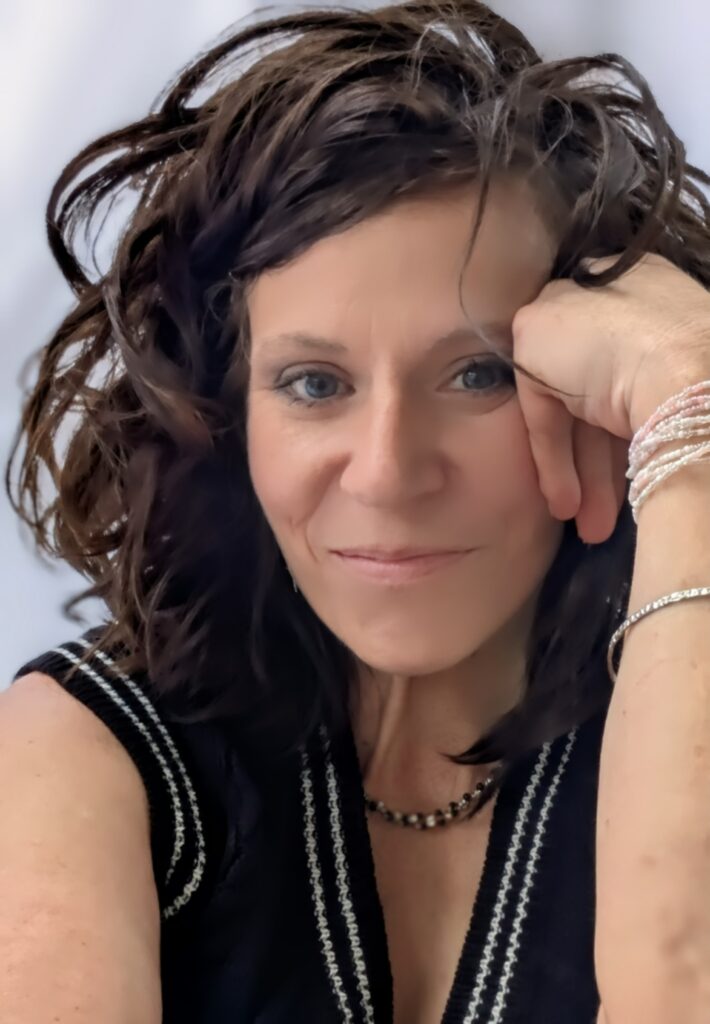 Picture of middle-aged dark hair woman with pale brown skin, curly hair, resting her face against her closed hand. Wearing a black knit sweater thats sleeveless and a dark beaded necklace.